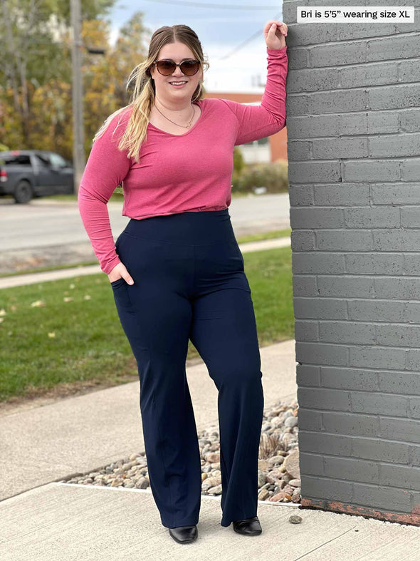 Miik model Bri (five feet five, xlarge) smiling next to a brick wall wearing Miik's Sierra high waisted pocket pant in navy with a pink long sleeve top tucked in