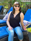 Miik model Sofia (five feet two, xsmall) sitting on a chair in the backyard wearing Miik's Skye ruffle neck blouse in black with jeans and sunglasses 