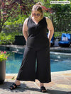 Miik model Bri (five feet five, xlarge) standing in front of a pool and looking down wearing an all black outfit: Miik's Skye ruffle neck blouse and a wide leg pant