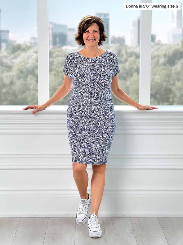 Woman standing in front of a window wearing Miik's Sofia reversible everyday dress in blue ditsy flower print.
