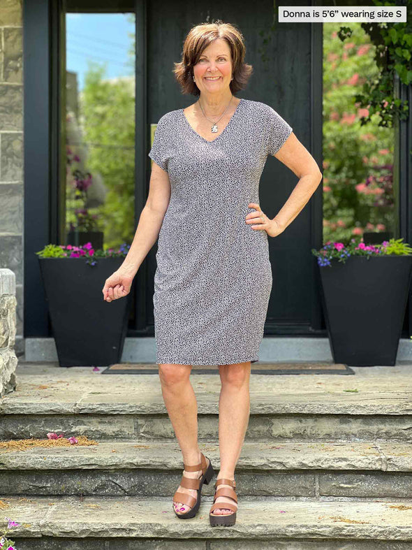 Miik model Christal (five feet three, large) smiling with one hand on her hips and the other holding a coat on her shoulders wearing Miik's Sofia reversible everyday dress in pebble print and black sandals 