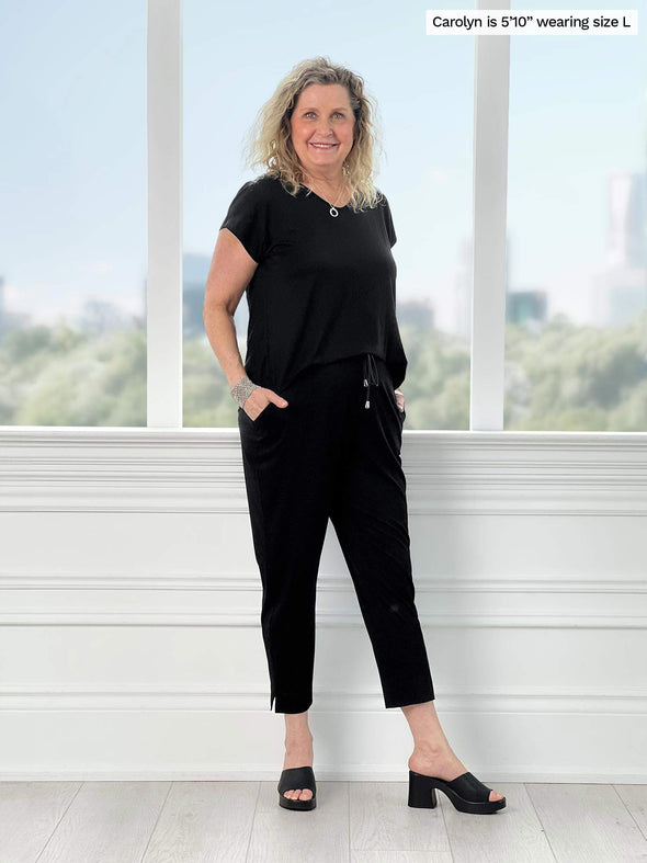 Miik model Carolyn (5'10", large) smiling while standing in front of a window wearing Miik's Stef v-neck open-back capri jumpsuit in black with black sandals 