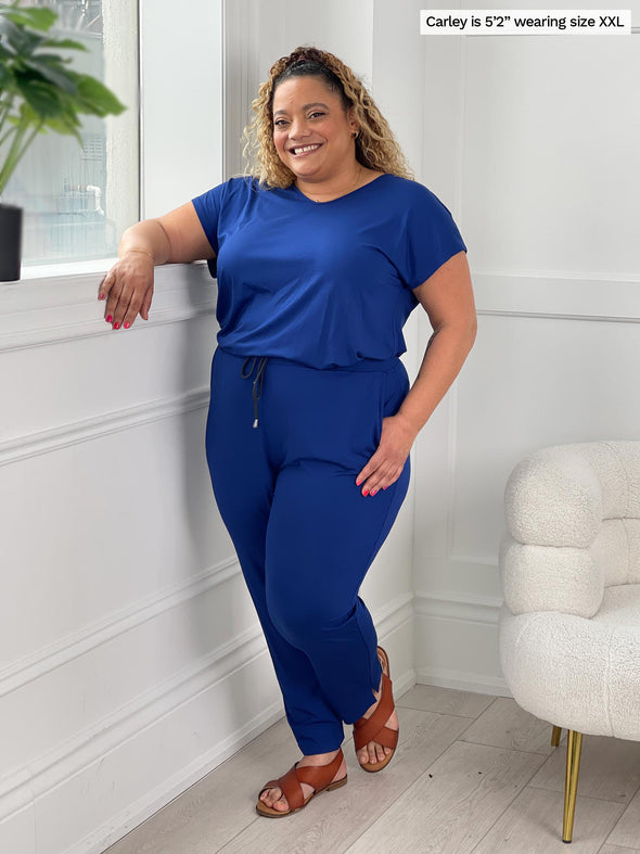 Miik model Carley (5'2", xxlarge) standing next to a white wall wearing Miik's Stef v-neck open-back capri jumpsuit in ink blue