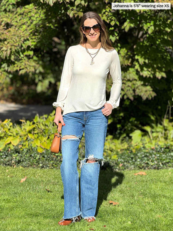 Miik model Johanna (5'6", xsmall) standing in nature smiling wearing a ripped jeans with Miik's Sunniva boatneck long sleeve top in natural, gold jewelry and sunglasses  