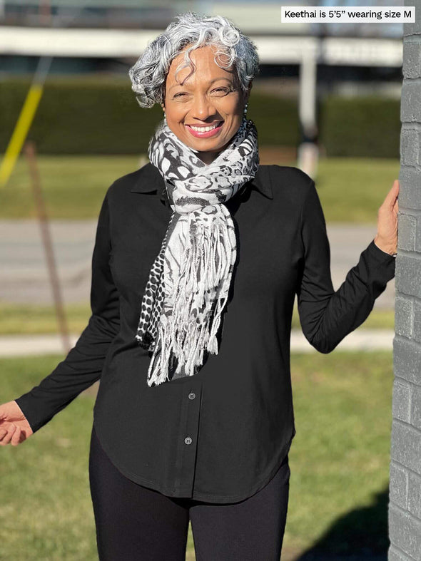 Miik model Keethai (5’5”, medium) smiling and standing next to a brick wall wearing Miik's Susan button up dress shirt in black with leggings and a animal print scarf in black and white