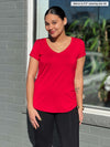 Miik model Meron (5'3", xsmall) smiling while standing in front of a window wearing Miik's Sutton v-neck classic tee in poppy red with a black pant 