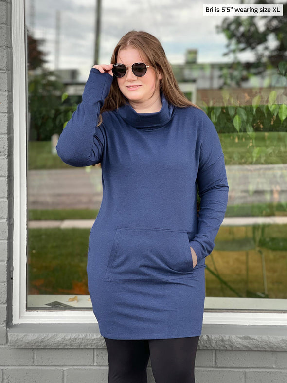 Woman standing in front of a window wearing Miik's Tate cowl neck lounge pocket tunic in navy with leggings.