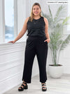 Miik model Christal (5'3", large) smiling while standing next to a window/white wall wearing Miik's Teanna high neck tank top in black wide pinstripe tucked in a tulip capri pant in black 