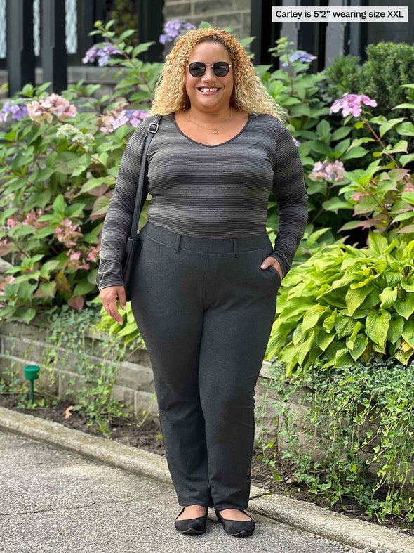 Woman sanding in front of plants wearing Miik's Tray long sleeve striped tee in charcoal stripe with charcoal pants. 