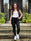Miik model Yasmine (5'0", xsmall, petite) smiling while standing in front of a door entry wearing Miik's Tyson mid-rise fleece legging in black with a tank top in white