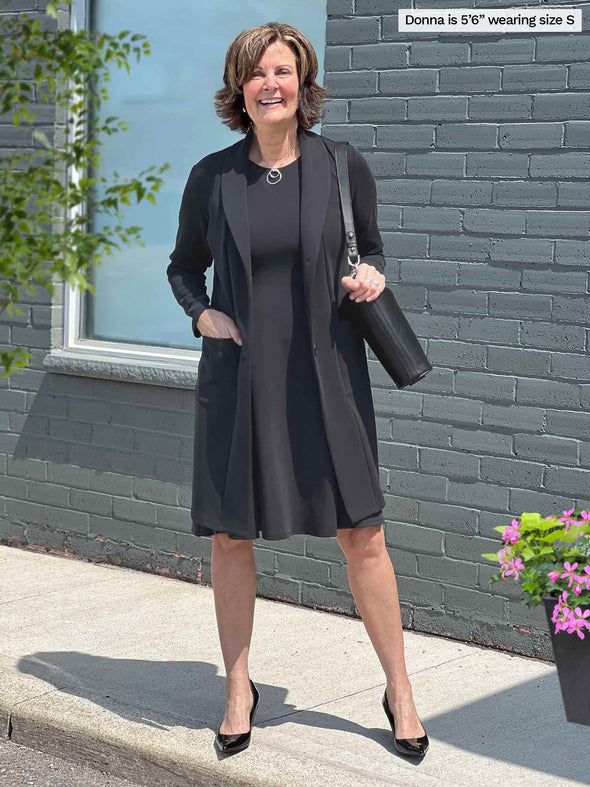 Miik founder Donna (five feet six, small) smiling standing in front of a brick wall wearing Miik's Valerie sleeveless fit and flare dress  in graphite along with a long coat in the same colour