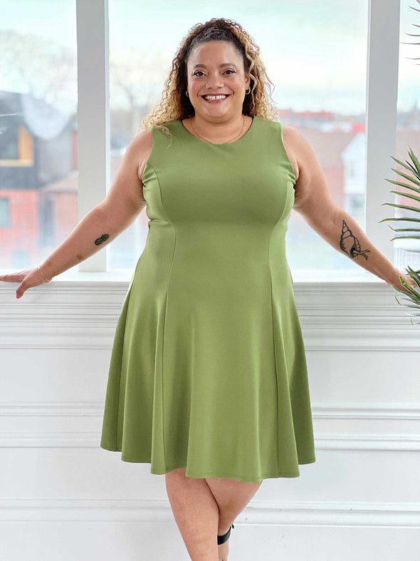 Miik model Carley (five feet two, size double extra large) smiling and standing in front of a window wearing Miik's Valerie sleeveless fit and flare dress in green moss