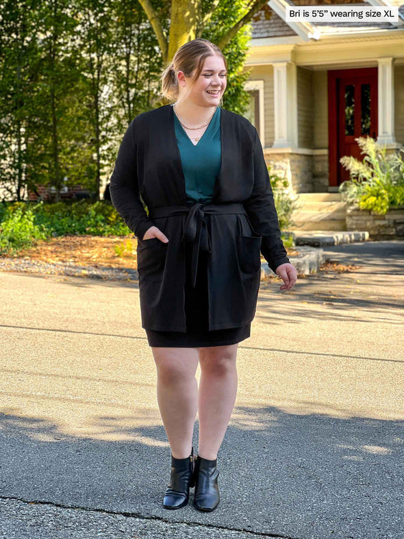 Miik model Bri (5’5”, xlarge) smiling while looking away wearing Miik's Vula belted cardigan with pockets in black with a matching colour pencil skirt, boots and a pine green v-neck shirt 