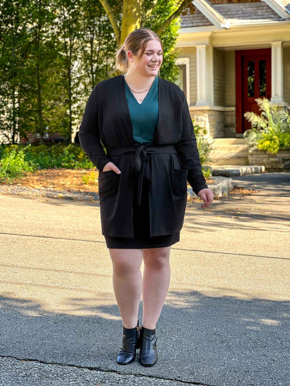 Miik model Bri (5’5”, xlarge) smiling while looking away wearing Miik's Vula belted cardigan with pockets in black with a matching colour pencil skirt, boots and a pine green v-neck shirt