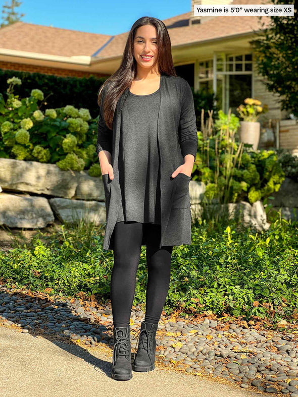 Miik model Yasmine (5'0", xsmall, petite) smiling with hands on pockets wearing Miik's Vula belted cardigan with pockets in charcoal with a tunic in the same colour