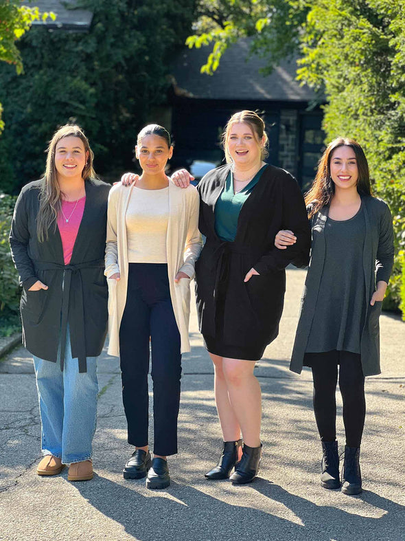 Miik models Christal, Meron, Bri and Yasmine smiling while standing next to each other showing the different colours and ways to wear the Miik's Vula belted cardigan with pockets. Colours available are: camel melange, charcoal and black