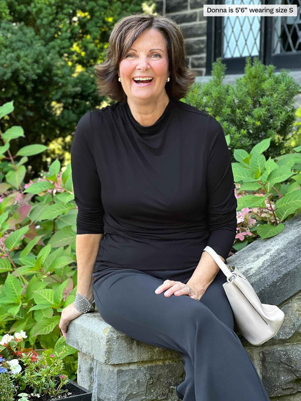 Miik founder Donna (five feet six, small) smiling while sitting in front of a garden wearing Miik's Wallace long sleeve draped blouse in black with a graphite flare pant 