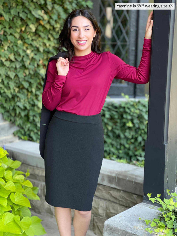 Miik model Yasmine (five feet tall, xsmall, petite) smiling wearing a pencil skirt in charcoal along with Miik's Wallace long sleeve draped blouse in bordeaux 