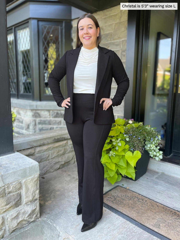 Miik model Christal (five feet three, large) smiling in front of a doorway wearing Miik's Wallace long sleeve draped blouse in natural tucked in a high waisted pocket pant in black with a blazer in the same colour 