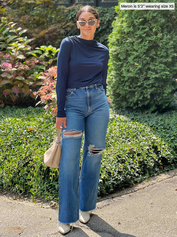 Miik model Meron (five feet three, xsmall) standing in front of a garden wearing Miik's Wallace long sleeve draped blouse in navy ticked in a ripped jeans