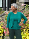 Miik model Keethai (5'5", medium) smiling wearing Miik's Wesley cropped cardigan in jade melange with a tank in the same colour and a flare pant in green pine