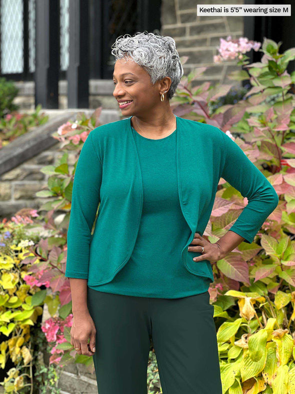 Miik model Keethai (5'5", medium) smiling wearing Miik's Wesley cropped cardigan in jade melange with a tank in the same colour and a flare pant in green pine