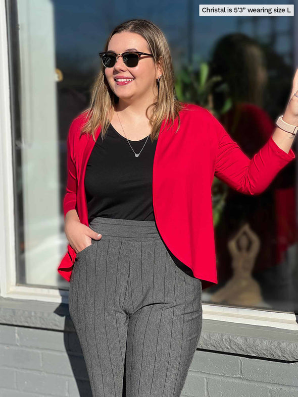 Woman standing in front of a window wearing Wesley cropped cardigan in poppy red over a black top and grey pants.