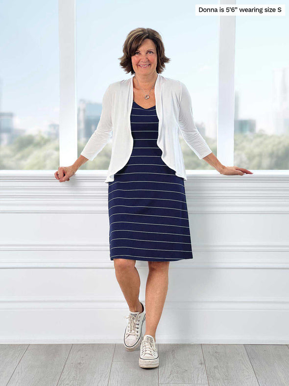 Miik founder Donna (56", small) standing in front of a window with arms open wearing a navy striped dress along with Miik's Wesley cropped cardigan in white 