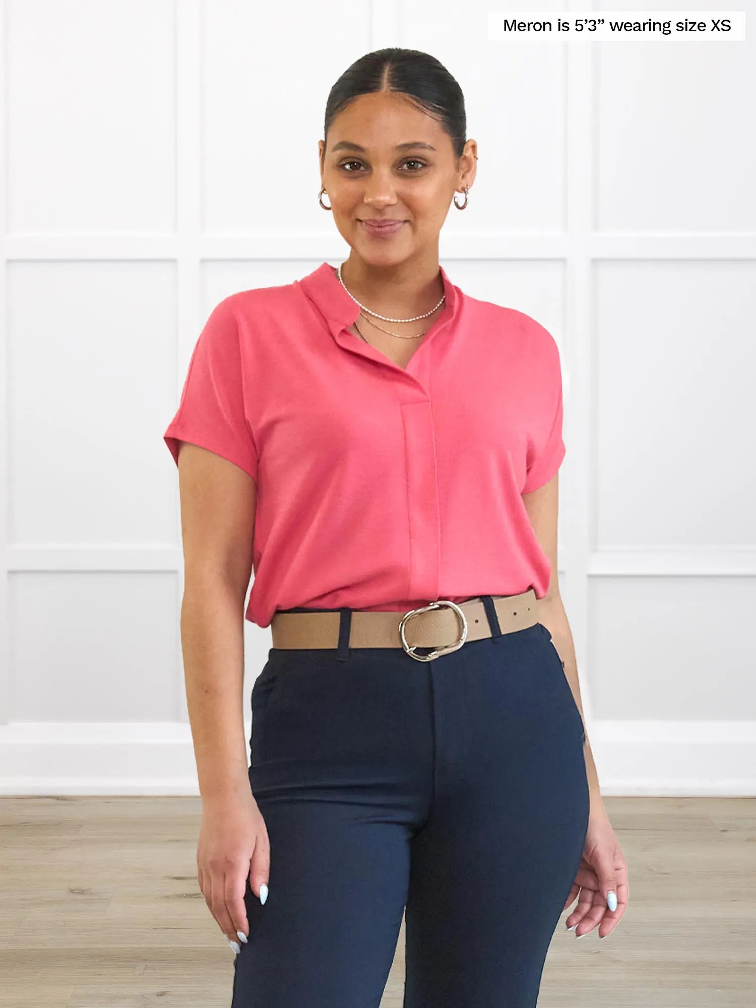Woman wearing a pink shirt and navy pants with a belt, standing against a white wall. 