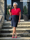 Miik model Keethai (5'5", medium) standing on backyard stairs wearing Miik's Yasmina band collar blouse in poppy red along with a midi skirt in navy pinstripe, sunglasses and a red sandals 