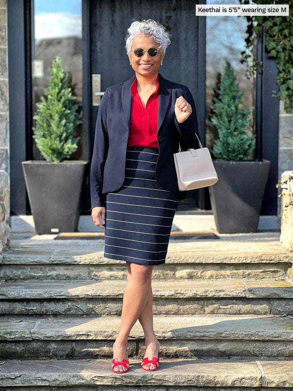 Miik model Keethai (5'5", medium) standing on backyard stairs wearing Miik's Yasmina band collar blouse in poppy red along with a midi skirt in navy pinstripe and a navy blazer 