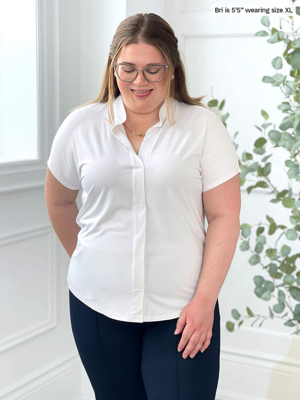 Miik model Bri (5'5", xlarge) smiling and looking down wearing Miik's Yasmina band collar blouse in white with navy pants