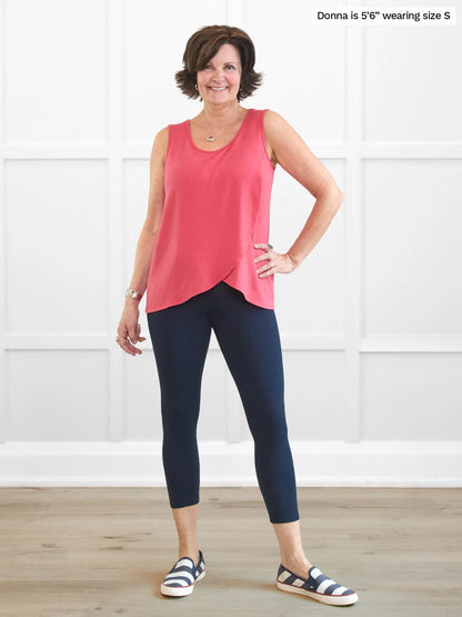Woman wearing a pink sleeveless top and navy leggings standing in a room with white paneled walls. 