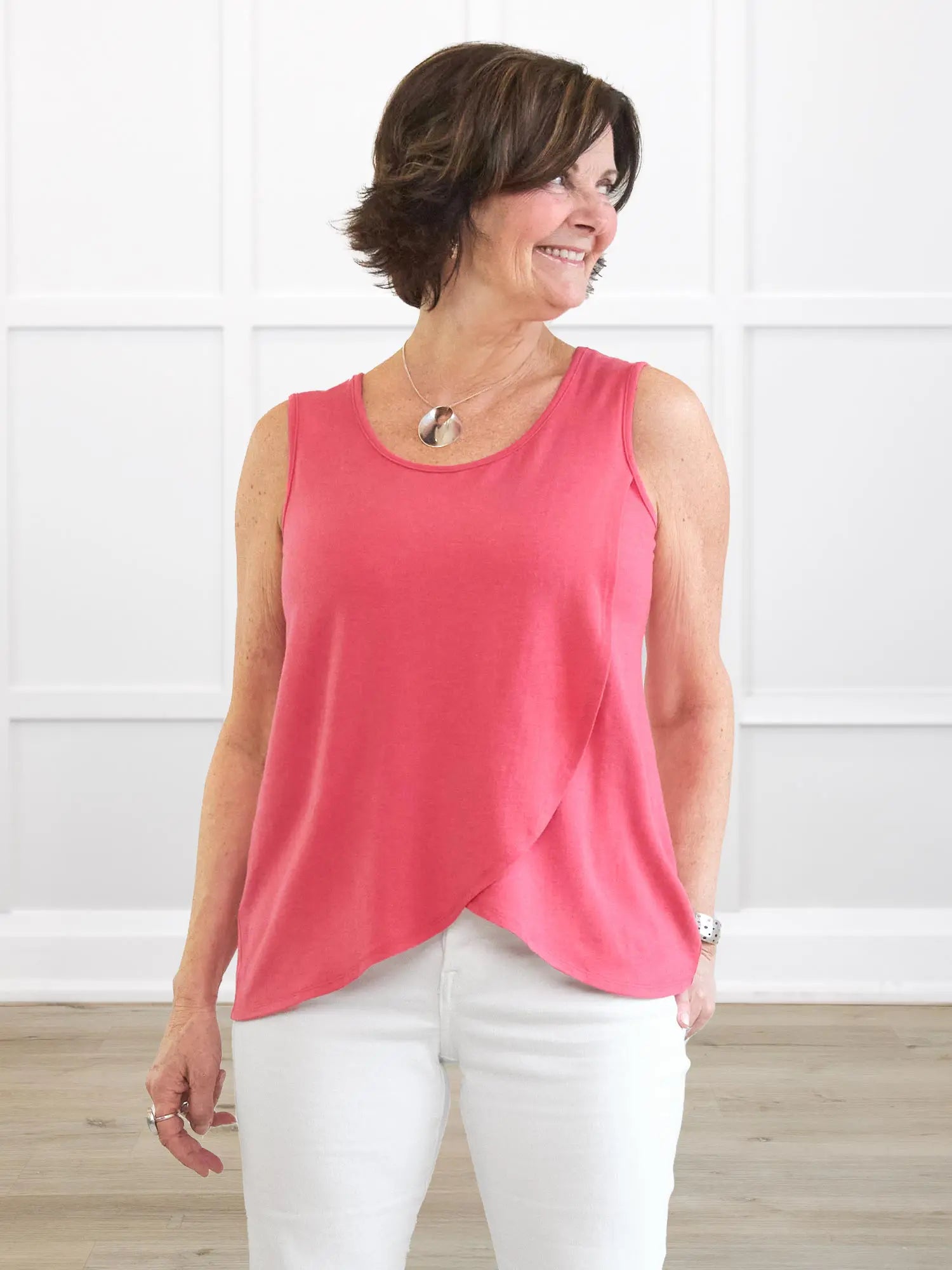 Woman wearing a pink sleeveless top and white pants against a white wall. 