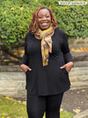 Miik model plus size Erica (5'7", size 2x) smiling with hands on pockets wearing Miik's Zuri long sleeve pocket tunic in black with a legging in the same colour and a yellow printed scarf