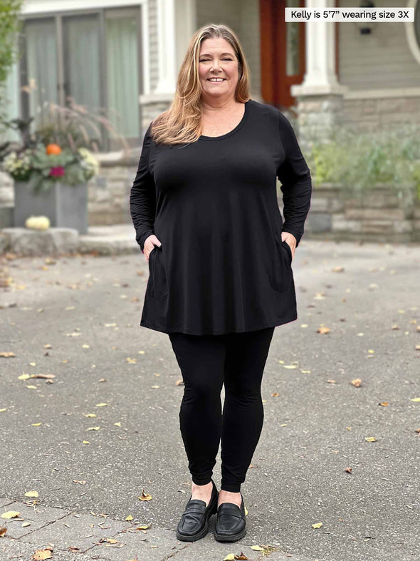 Miik model plus size Kelly (5'7", size 3x) smiling wearing an all black outfit: Miik's Zuri long sleeve pocket tunic and Lisa2 legging