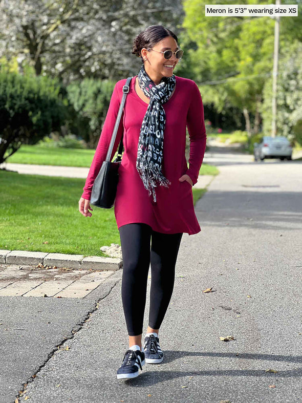 Miik model Meron (five feet three, xsmall) walking with hands on her pockets wearing  Miik's Zuri long sleeve pocket tunic in bordeaux with a black legging and a scarf