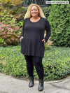 Miik model Carley (five feet two, xxlarge) smiling while standing in front of garden with hands on her pockets wearing Miik's Zuri long sleeve pocket tunic in charcoal, black legging and boots