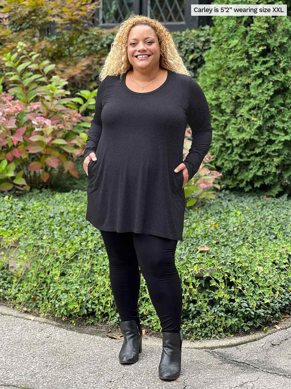 Miik model Carley (five feet two, xxlarge) smiling while standing in front of garden with hands on her pockets wearing Miik's Zuri long sleeve pocket tunic in charcoal, black legging and boots