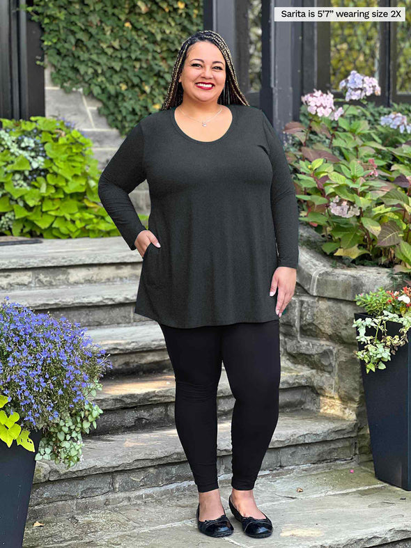 Miik model plus size Sarita (5'7", 2x) smiling while standing on outdoor stairs wearing Miik's Zuri long sleeve pocket tunic in charcoal with a black legging