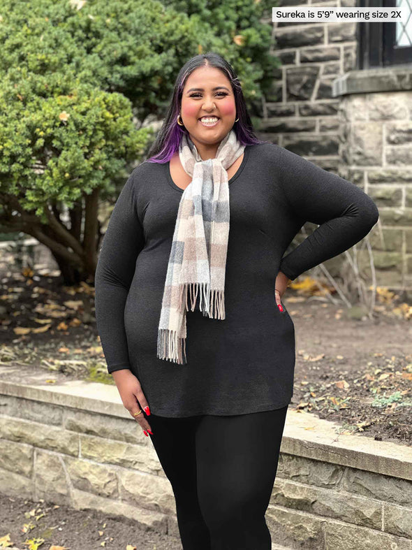 Miik model plus size Sureka (5'9", size 2x) smiling while standing in front of a house wearing Miik's Zuri long sleeve pocket tunic in charcoal with a black legging and a neutral coloured scarf 