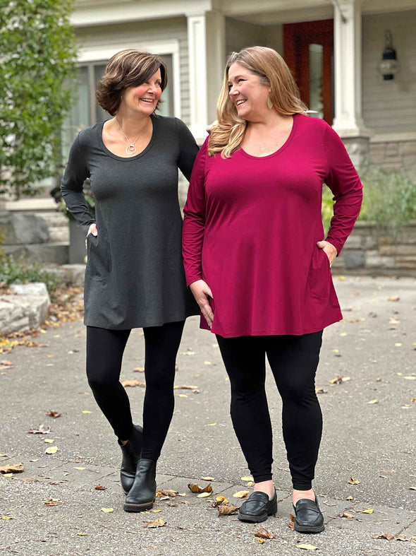 Miik founder Donna and model Kelly smiling while looking at each other both wearing the same outfit but in different colours. They are both wearing high waited legging in black with Miik's Zuri long sleeve pocket tunic in charcoal and bordeaux
