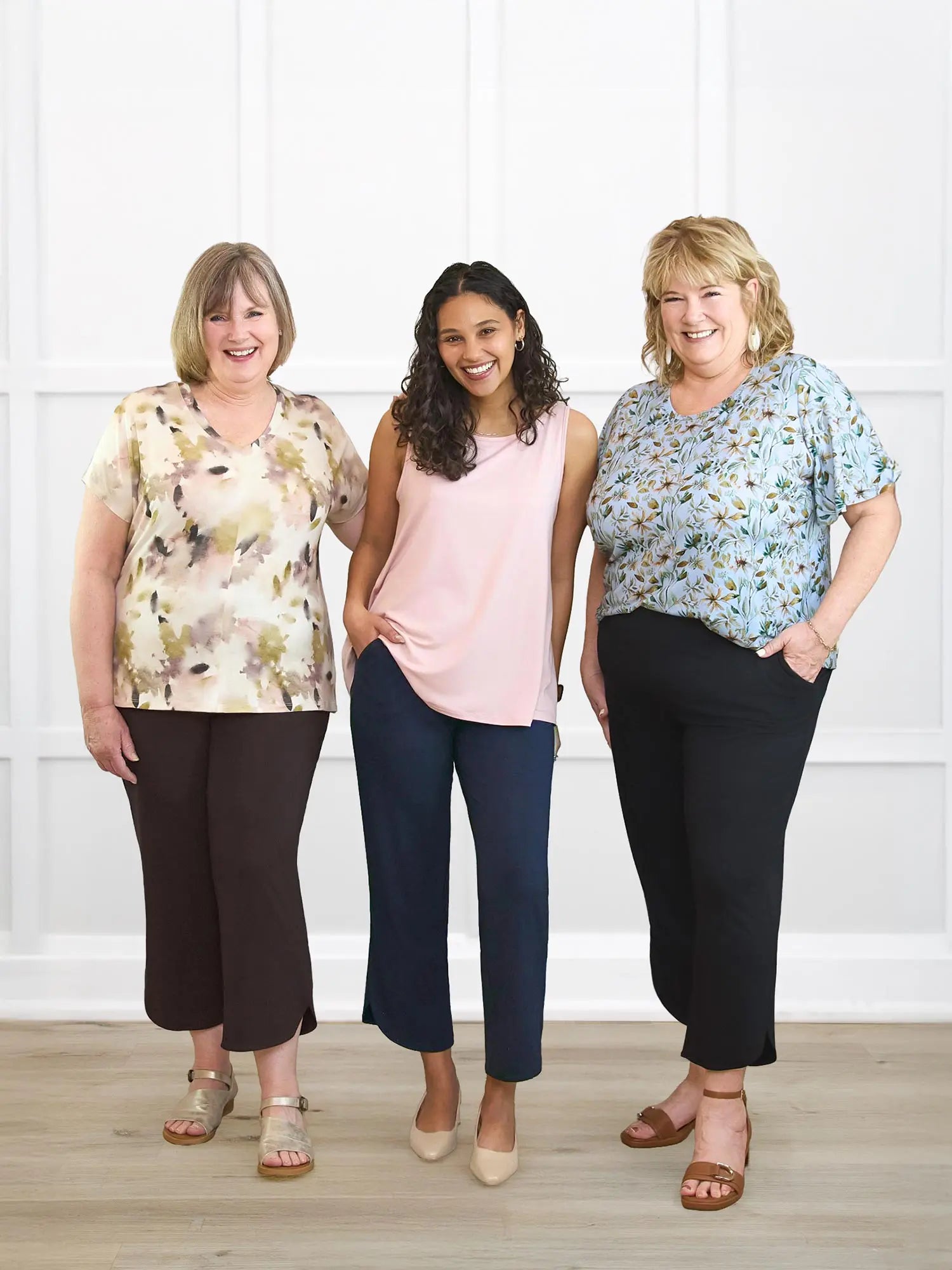 Three women standing together wearing floral and plain tops with dark pants against a white wall.