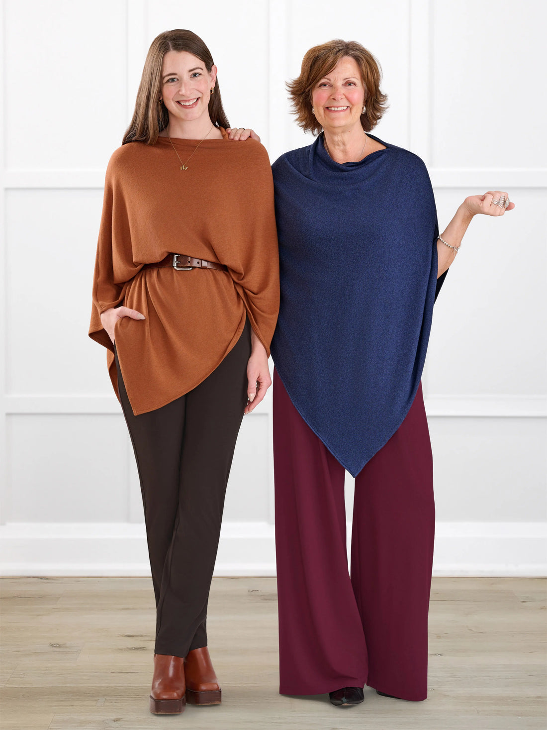 Two women modeling draped poncho-style tops in a bright indoor studio setting. The woman on the left wears a rust brown poncho with dark brown pants and brown shoes.