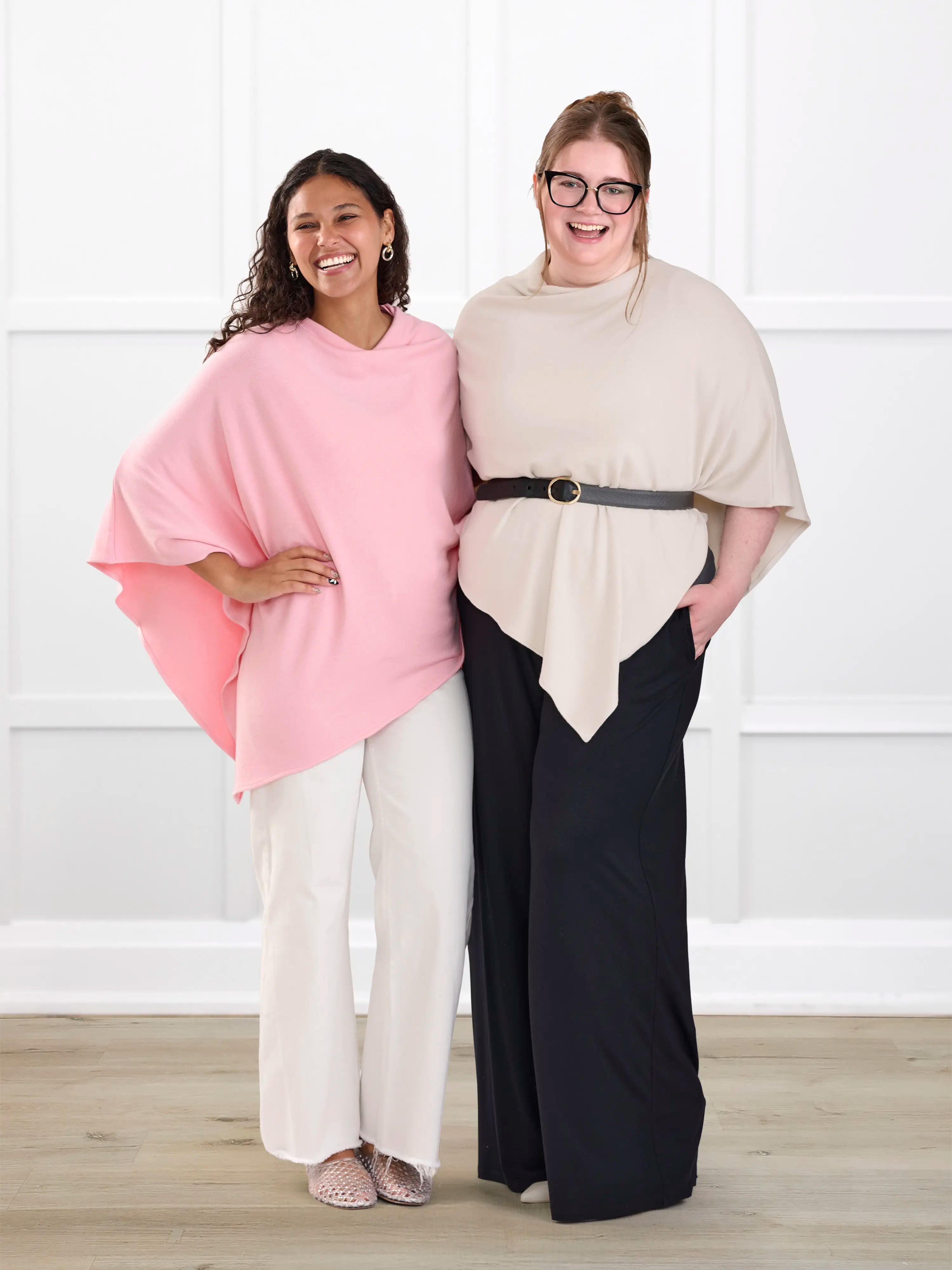 Two women modeling draped poncho-style tops in a bright indoor studio setting. The woman on the left wears a light pink top with white wide-leg pants, and the woman on the right wears a beige top with a black belt and black wide-leg pants.