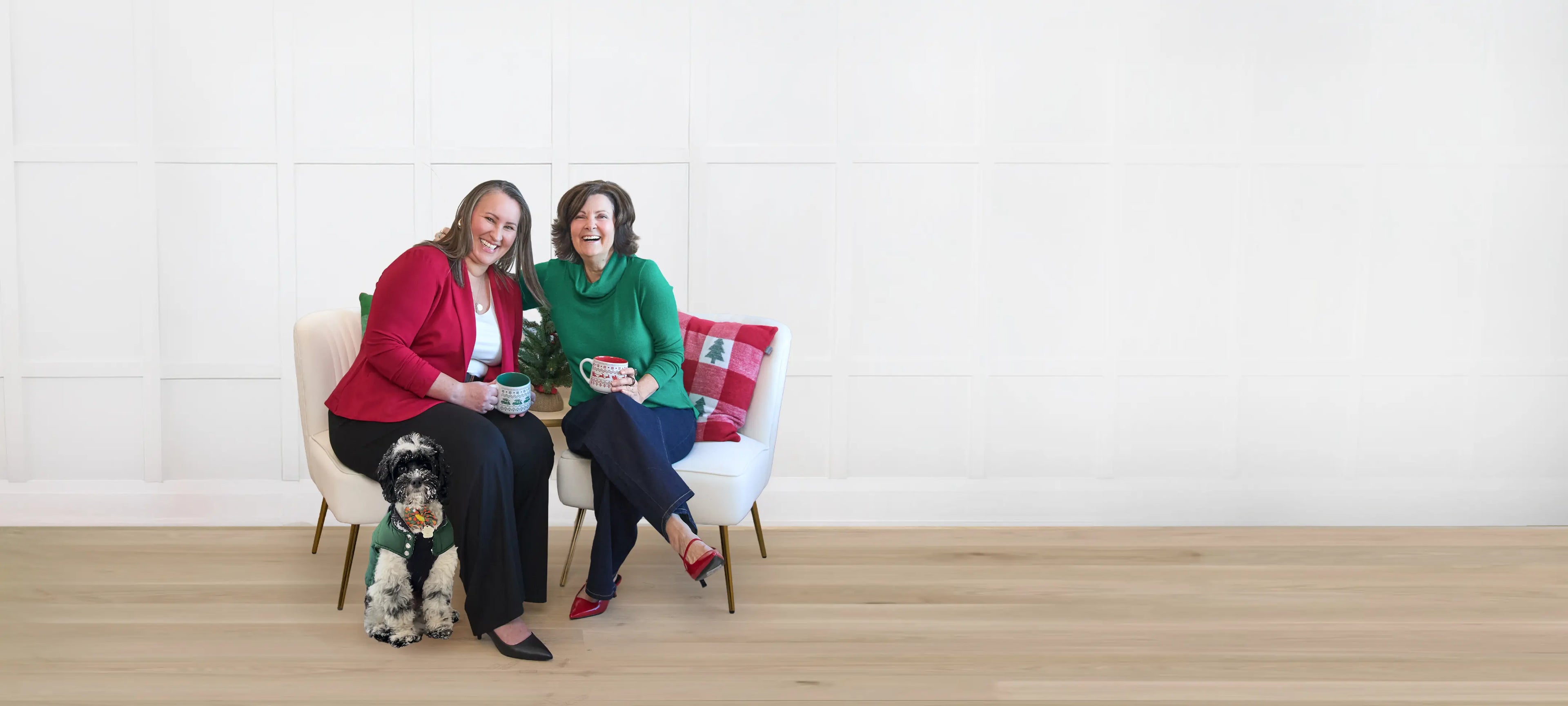 Two women sitting on a white couch holding mugs with a small black and white dog wearing a sweater on the floor next to them