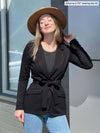 Woman standing in front of a window looking up with closed eyes wearing a black blazer belted with Miik's Albany reversible long sleeve tee in charcoal/granite melange, jeans and a brown hat