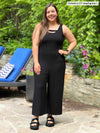Woman standing in front of patio lounge chairs smiling while wearing Miik's Alice reversible capri wide leg jumpsuit in black.