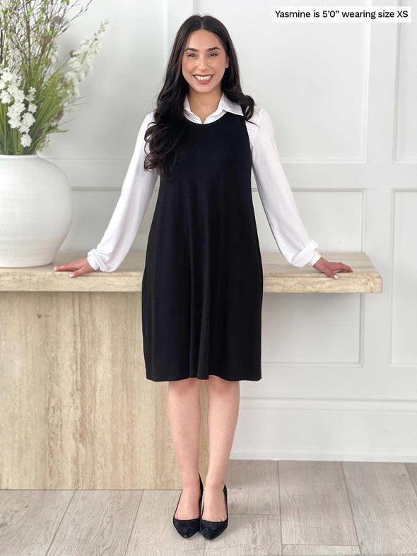 Miik model Yasmine (five feet. tall, size extra small, petite) smiling standing in front of white wall wearing a collared white top underneath of Miik's Aubrey reversible swing dress in black
