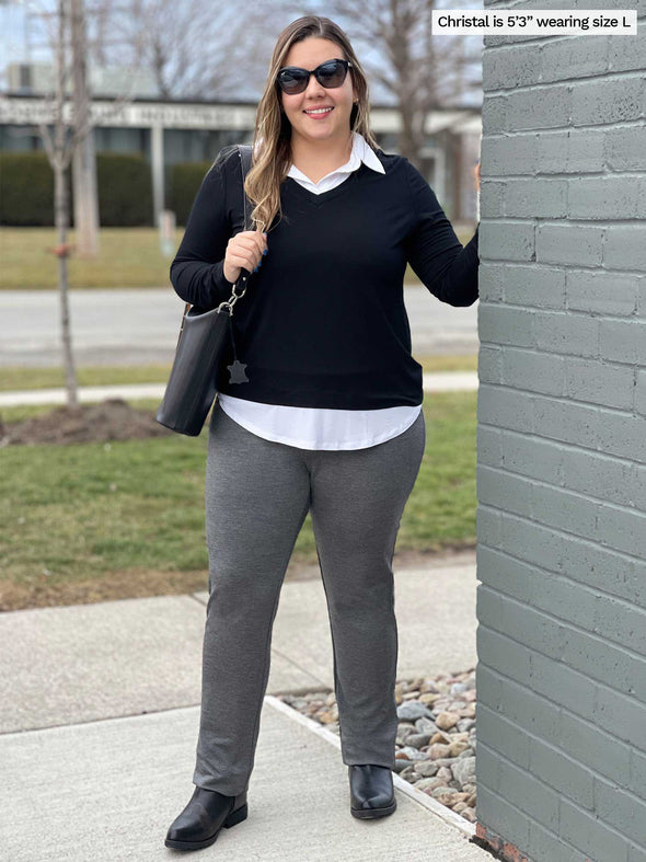 Woman standing next to a brick wall and smiling wearing Miik's Avery pull-on pant in granite melange french terry along with a long sleeve collared faux-layer shirt in black and white, sunglasses and a black purse on her shoulders 